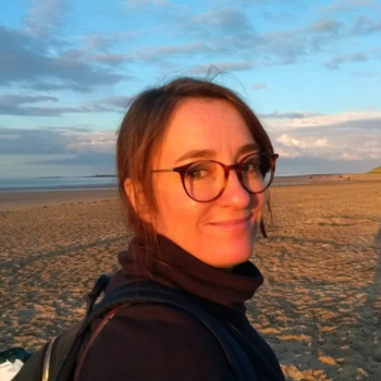 Woman wearing glasses on beach at sunset, looking toward camera with backpack.